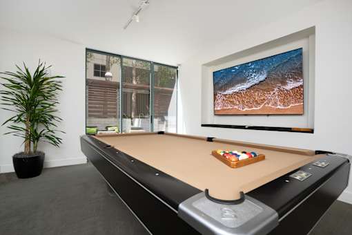 A pool table with a brown cloth and a television on the wall.