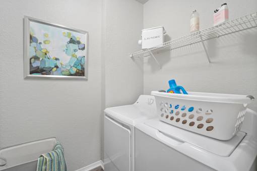 A laundry room with a white washer and dryer, a basket on top of the dryer, and a painting on the wall at The Grand Reserve at Tampa Palms Apartments, Tampa, FL, 33647