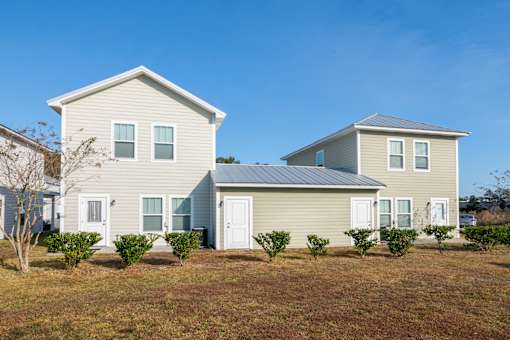 A house with a grey roof and white trim is surrounded by bushes and has a clear blue sky in the background.
