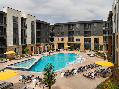 A large swimming pool surrounded by lounge chairs and umbrellas in front of apartment buildings.
