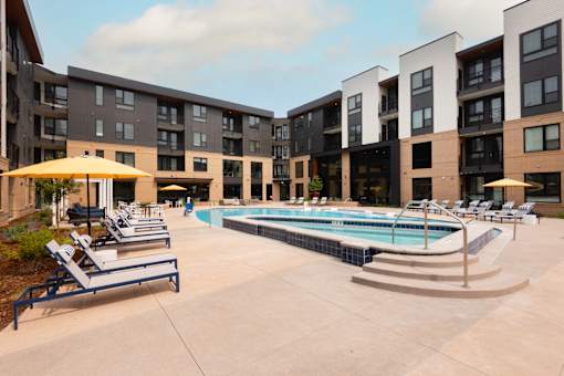 A swimming pool surrounded by sun loungers and umbrellas in front of apartment buildings.