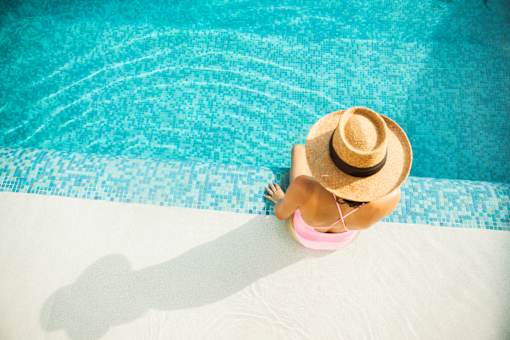 A woman in a straw hat and pink bikini sits by a pool.