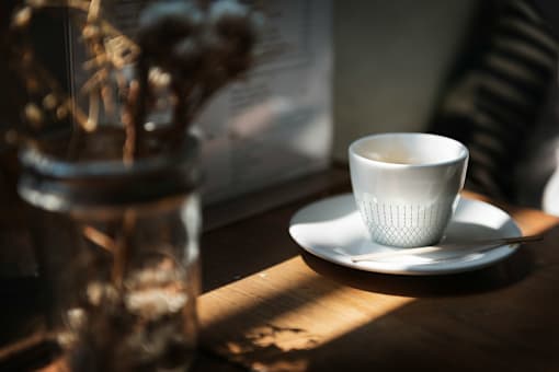 A white cup sits on a saucer with a vase of dried flowers in the background.