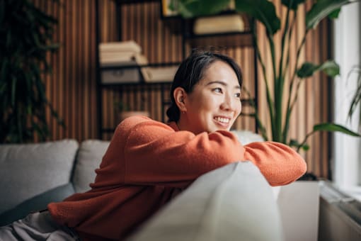 A woman in an orange top is sitting on a grey couch.