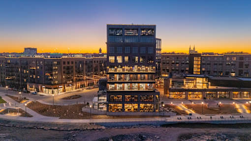 A modern building with a glass facade is illuminated from within, set against a twilight sky.