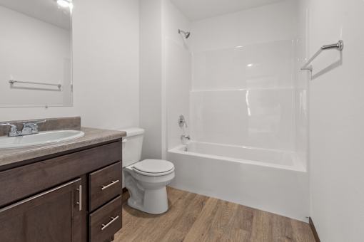 A white toilet sits in a bathroom with a wooden floor and a white tub.