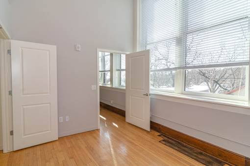A room with white walls and wooden floors has two white doors and a window with blinds.