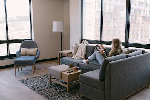 A woman is sitting on a grey couch in a living room with a large window behind her.