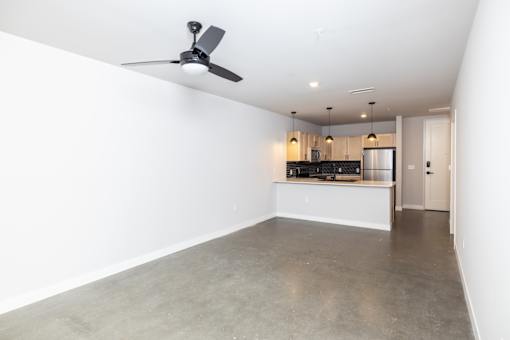 A room with a ceiling fan and a kitchen area in the background.