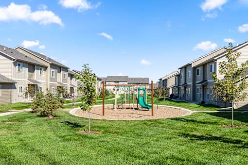 A playground with a green slide in the middle of a grassy area surrounded by houses.