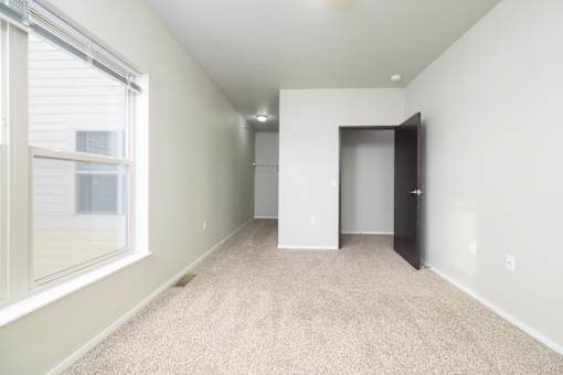 A room with a carpeted floor, a window with blinds, and a door leading to another room.
