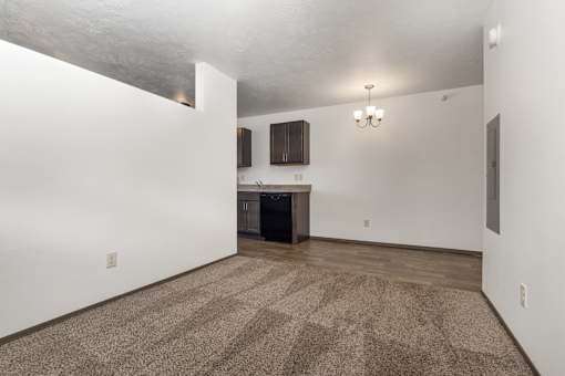 A room with a carpeted floor and a kitchen area in the background.