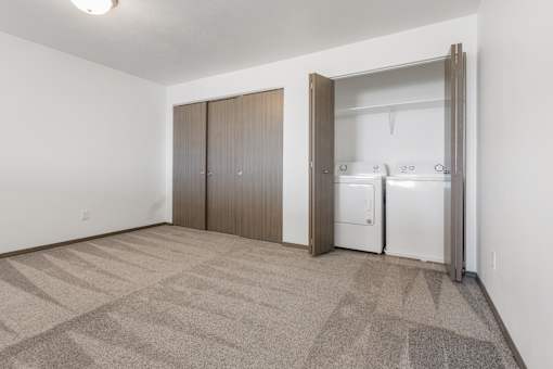 A room with a carpeted floor and a white wall with a fridge and cupboards in it.