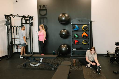 A woman is sitting on a mat in a gym, stretching her legs.