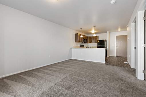 A spacious living room with a carpeted floor and a kitchen area in the background.