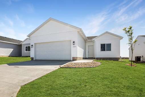 A white house with a grey garage door and a small tree in front.