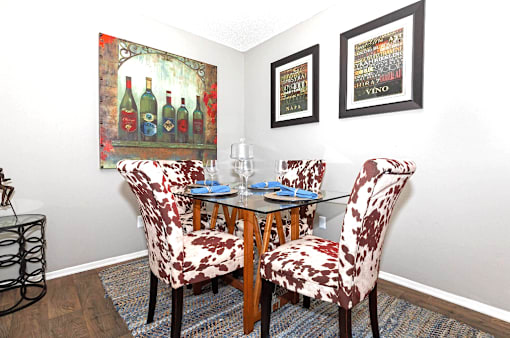 A dining room with a glass table and chairs with a wine bottle painting on the wall.