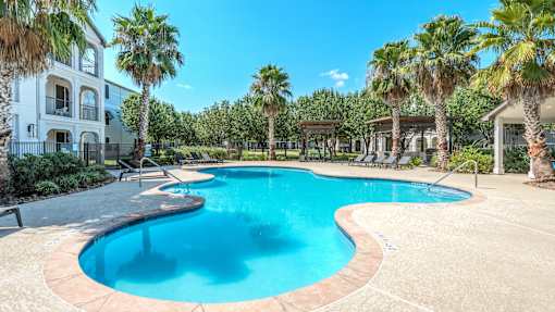 A large swimming pool surrounded by palm trees and a white building at Rosemary Apartments, Conroe, Texas