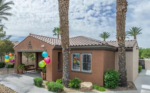 A house with a brown roof and tan walls is surrounded by palm trees and has balloons tied to the front door.
