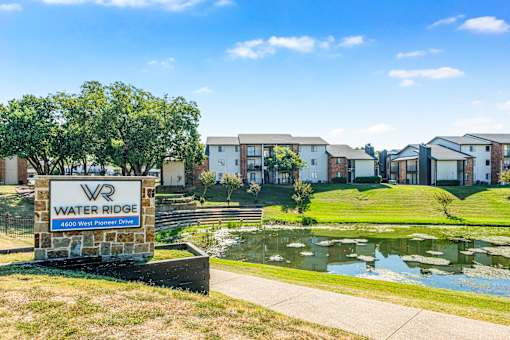 A sign for Water Ridge sits in front of a pond and houses at Water Ridge Apartments , Irving, TX
