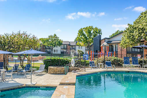 A swimming pool surrounded by lounge chairs and umbrellas with a building in the background at Water Ridge Apartments , Irving, TX
