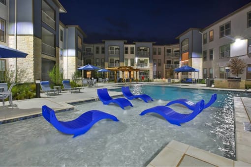 an outdoor pool with blue chairs and an apartment building at night at The Grand at Stonecreek, San Marcos, 78666