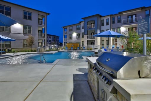 a grilling station next to a swimming pool with an apartment building in the background at The Grand at Stonecreek, San Marcos, TX, 78666