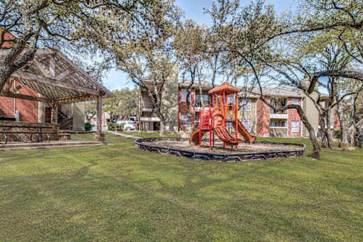 A playground with a red swing set is surrounded by trees and a building in the background.
