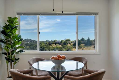 A table with a bowl of fruit on it sits in front of a window with a view of the outdoors.