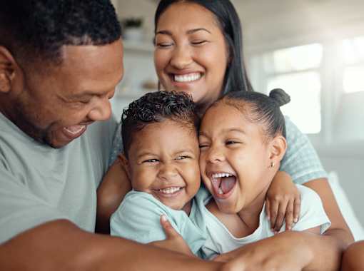 A family of four is hugging and smiling together in a bright room.