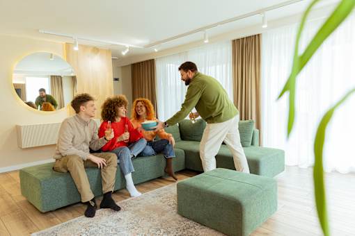 A man is serving food to two people sitting on a green couch in a bright room.