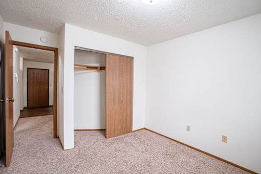 A room with a carpeted floor and a white wall with a door and a closet. Fargo, ND Cedars 4 Apartments