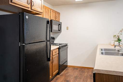 A black refrigerator stands in a kitchen with wooden cabinets and a stainless steel sink. Fargo, ND Cedars 4 Apartments