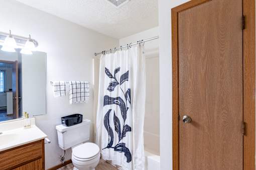 A bathroom with a toilet, sink, and shower curtain. Fargo, ND Candlelight Apartments