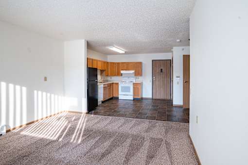A sunlit living room with a carpeted floor and a kitchen area in the background. Dilworth, MN Fieldstone Apartments