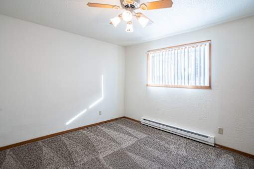 A room with a ceiling fan, a window with blinds, and a carpeted floor. Fargo, ND Kennedy Apartments