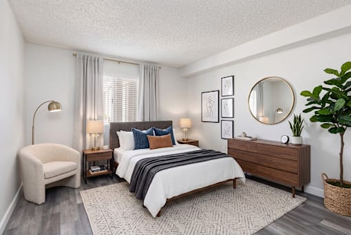 A bedroom with a large bed, a chair, a side table with lamps, a dresser, and a potted plant.