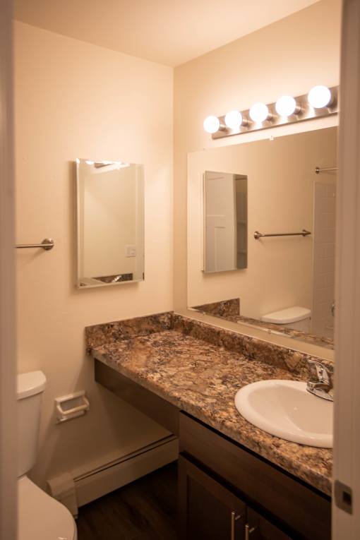 A bathroom with a granite counter top and a mirror with lights above it.