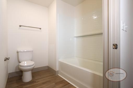 A bathroom with a white toilet and tub with shower and hardwood vinyl flooring.