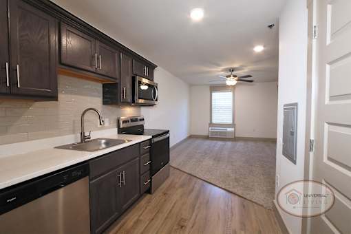 A view of a one bedroom apartment home with dark cabinets, white countertops and walls, carpet in the living area and hardwood vinyl flooring in the kitchen.