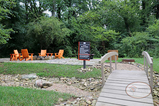 A walkway leads to a wooden bridge and a fire pit area with picnic table and lounge seating.