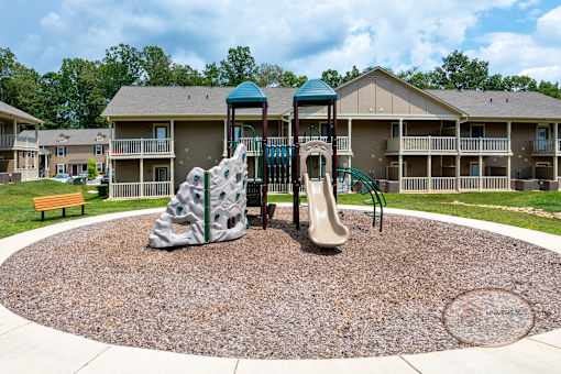 A playground with a slide, rock climber, and a wooden bench in front of apartment buildings.