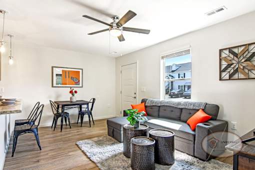 An open window lets light into a white living room with a grey couch, a dining table with chairs, and a ceiling fan.
