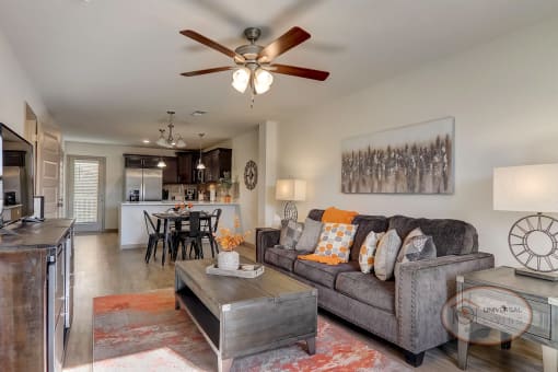 A living room with white walls, vinyl hardwood flooring, and dark cabinets. Furnished with a brown leather couch, grey armchair, orange dining table, a coffee table, flat screen TV, and plenty of orange decor.