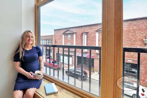 A woman sitting in the windowsill of a large window in a living room with white walls,  a brick building and downtown street can be seen outside.