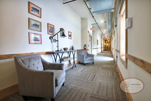 A hallway with a sitting area complete with table, lamp, two grey armchairs, and artwork.