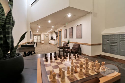 A close up of a chess board sitting on a table with a lounge in the background, complete with seating and mail center.