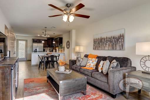 A fully furnished living room with hardwood vinyl flooring, white walls, and a kitchen in the background.