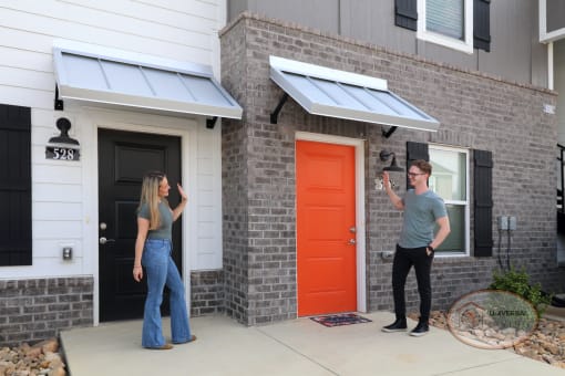 A man and a woman, each standing in front of the door to their apartment building, waving at one another.