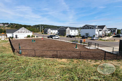 A landscape view of the fenced in dog park with obstacles, bench, and a water fountain for pets.